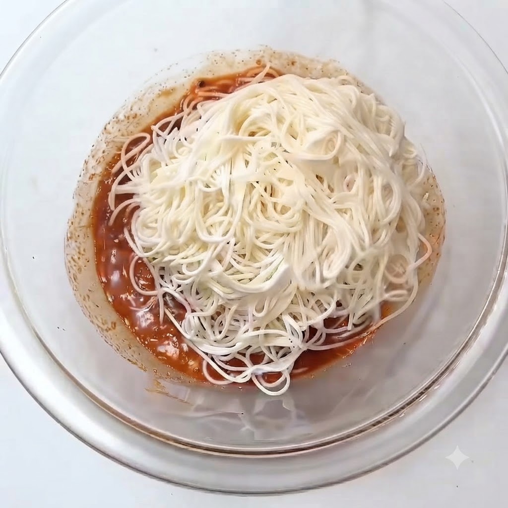 Un montón de fideos somen cocidos blancos colocados sobre la salsa rojiza dentro de un bol de cristal.