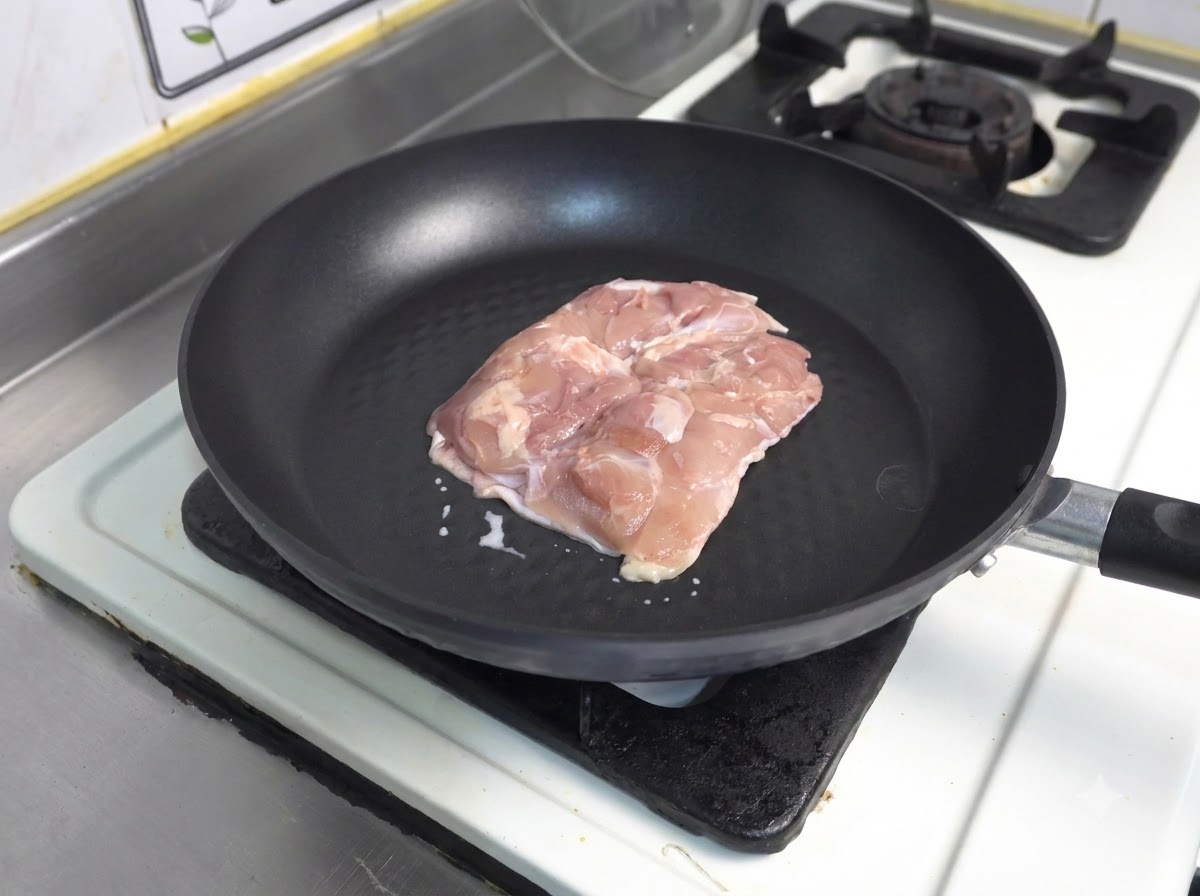 Un trozo de muslo de pollo crudo con la piel hacia abajo en una sartén antiadherente oscura.