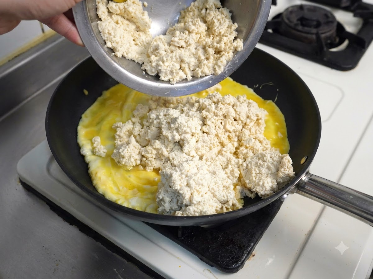 Un bol metálico vertiendo tofu blanco desmenuzado sobre huevos revueltos amarillos cocinándose en una sartén negra.