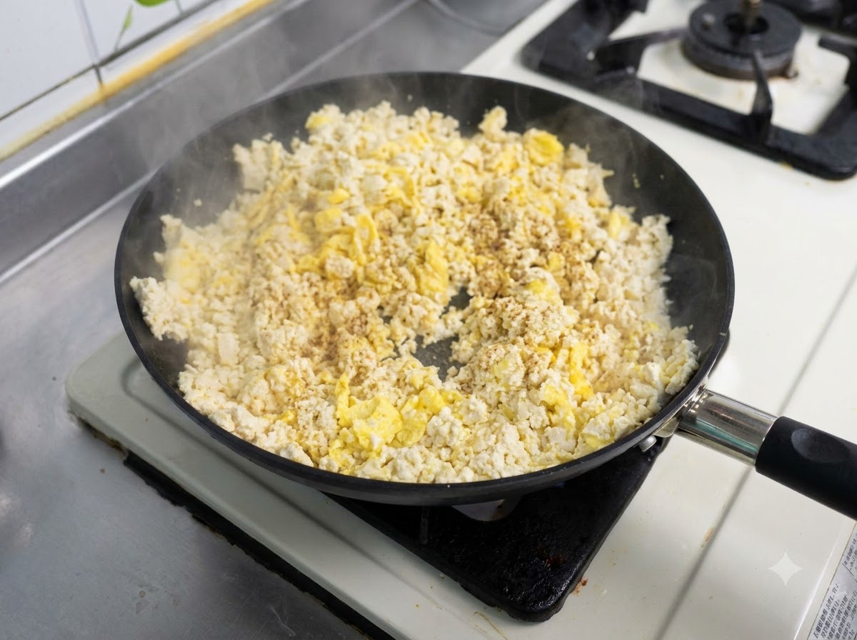 Una sartén llena con una mezcla de huevos revueltos y tofu siendo salteada con sazonador espolvoreado encima.