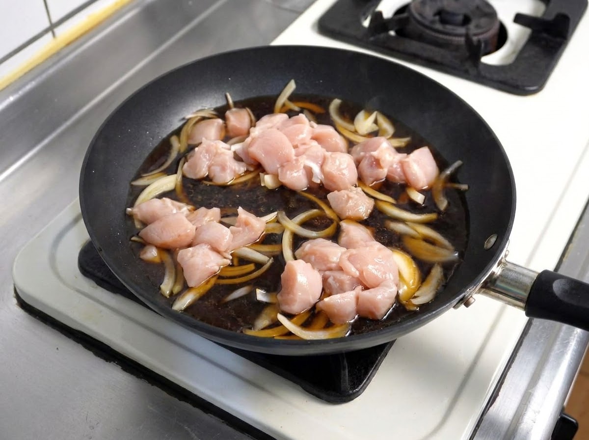 Trozos de pollo crudo colocados en una salsa oscura a fuego lento con cebollas dentro de una sartén.