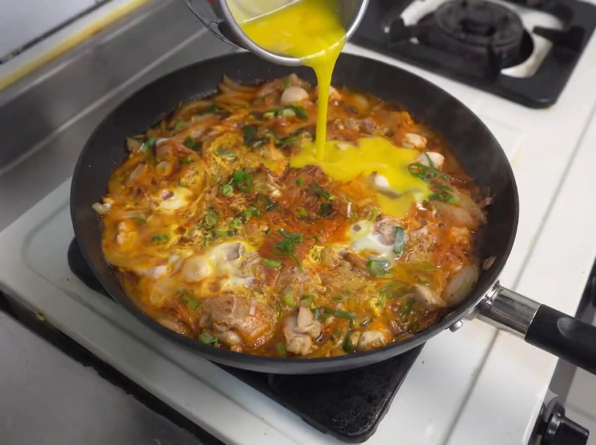 Vertiendo más huevos batidos desde un recipiente hacia la sartén a fuego lento con pollo y kimchi.