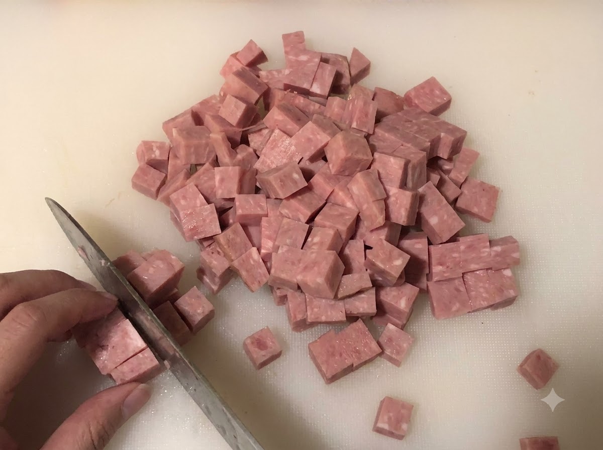 Spam cortado en cubitos pequeños sobre una tabla de cortar blanca con cebolla y cebollino picados al fondo.