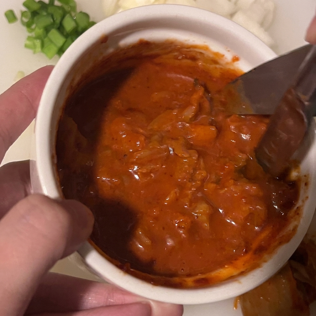 Tijeras de cocina cortando trozos de kimchi sumergidos en una salsa roja espesa dentro de un cuenco blanco pequeño.