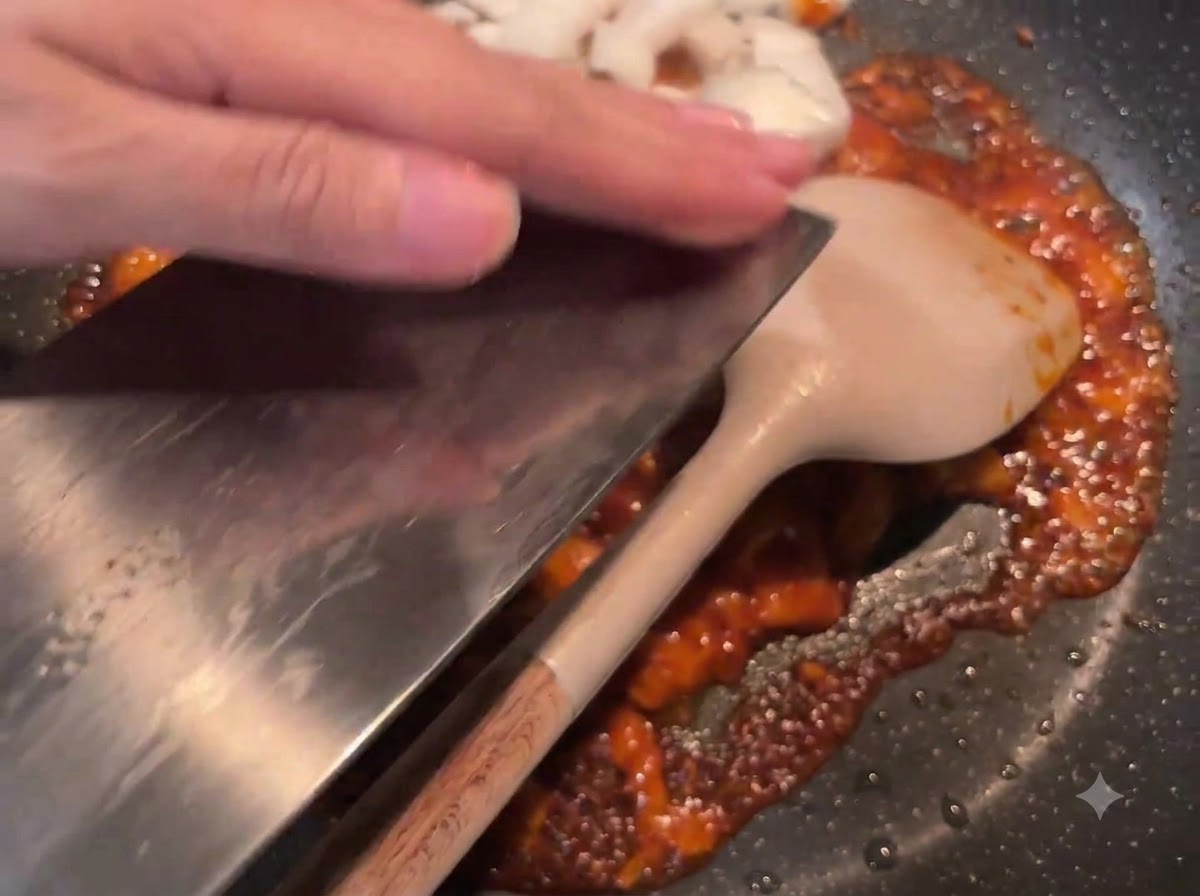 Cebolla blanca en dados siendo deslizada desde un cuchillo hacia la sartén con la salsa roja salteada.