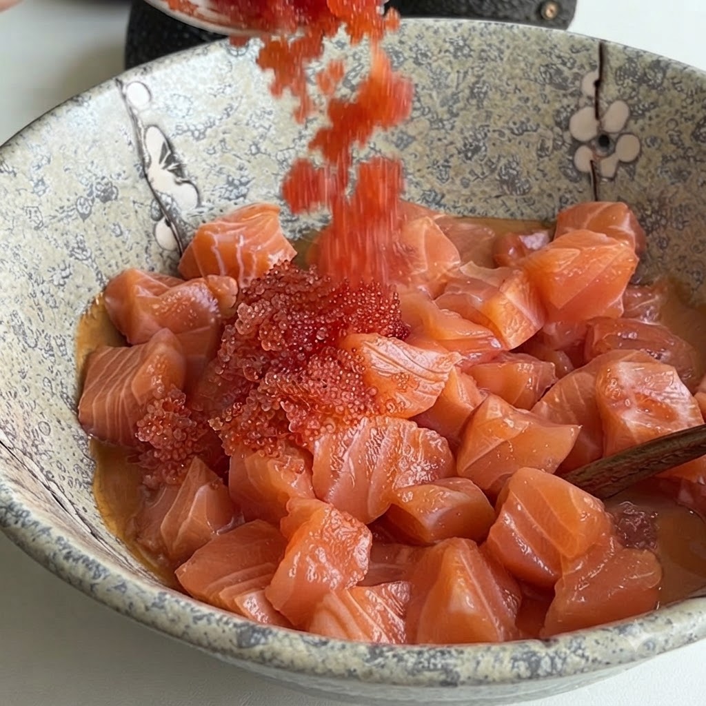 Hueva de pescado de color rojo brillante siendo vertida desde una cuchara en un tazón de cerámica lleno de salmón en cubos y salsa.