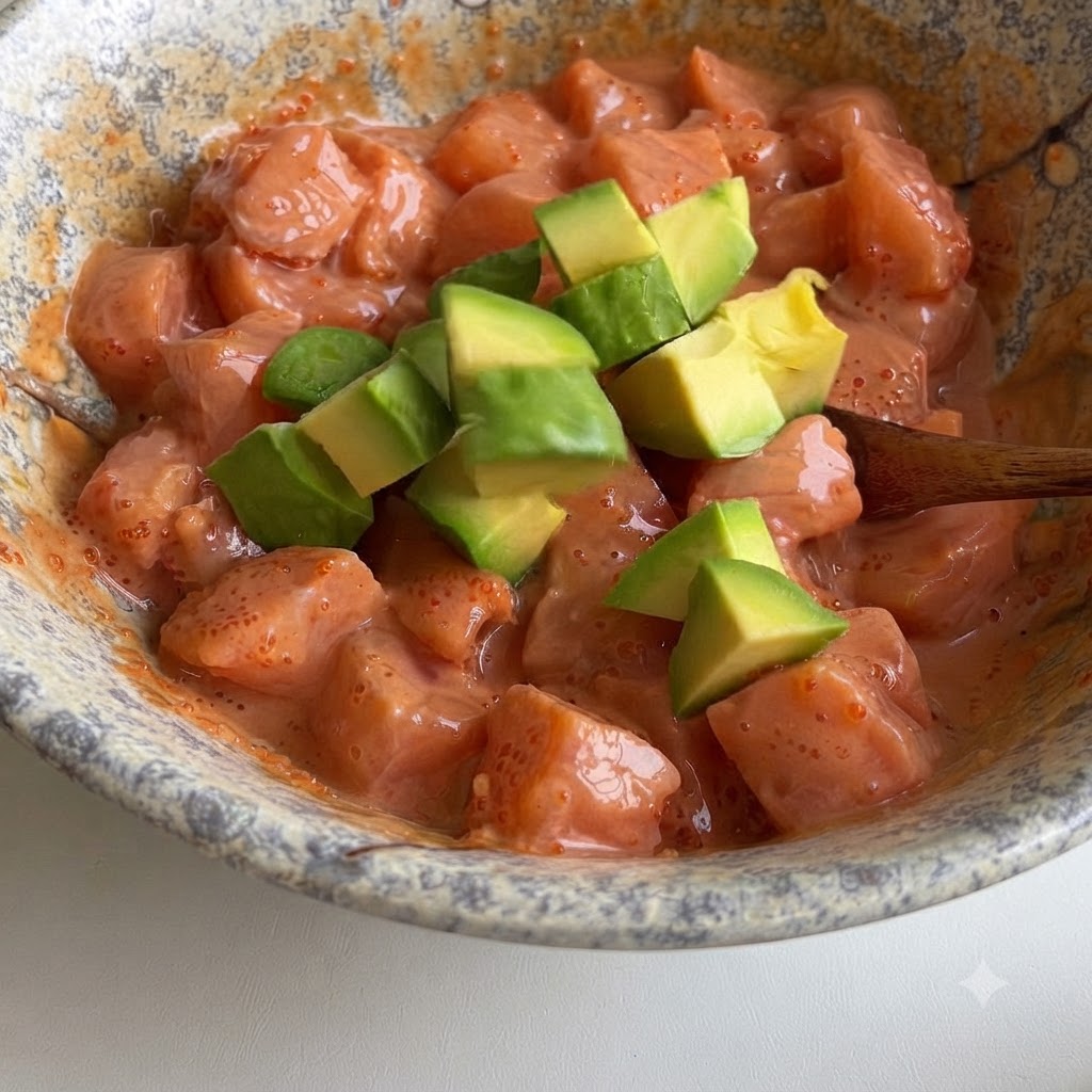Cubos de aguacate verde siendo doblados suavemente en un tazón de cubos de salmón cubiertos con una salsa rosa cremosa.