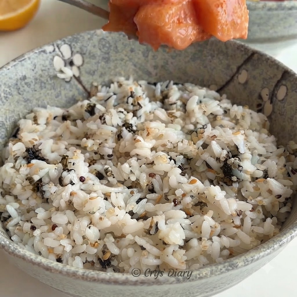 Una cuchara de mezcla de salmón siendo colocada sobre un tazón de cerámica lleno de arroz blanco y hojuelas de alga.