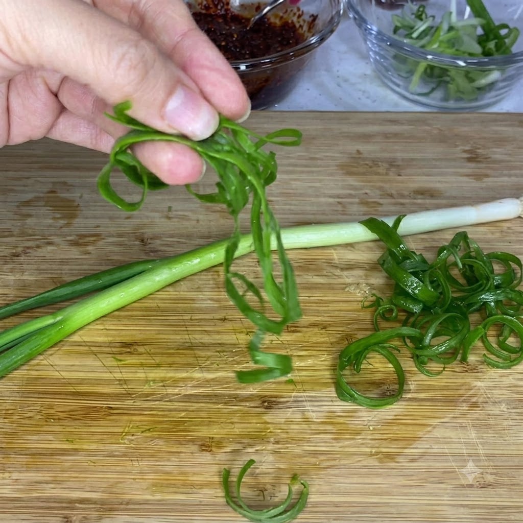 Una mano sosteniendo tiras de cebollina rizadas sobre una tabla de cortar de madera, con boles de vidrio con salsa y cebollas en rodajas al fondo.