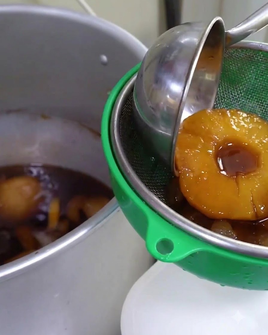Salsa infusionada con frutas siendo colada a través de un colador verde en un recipiente metálico grande.