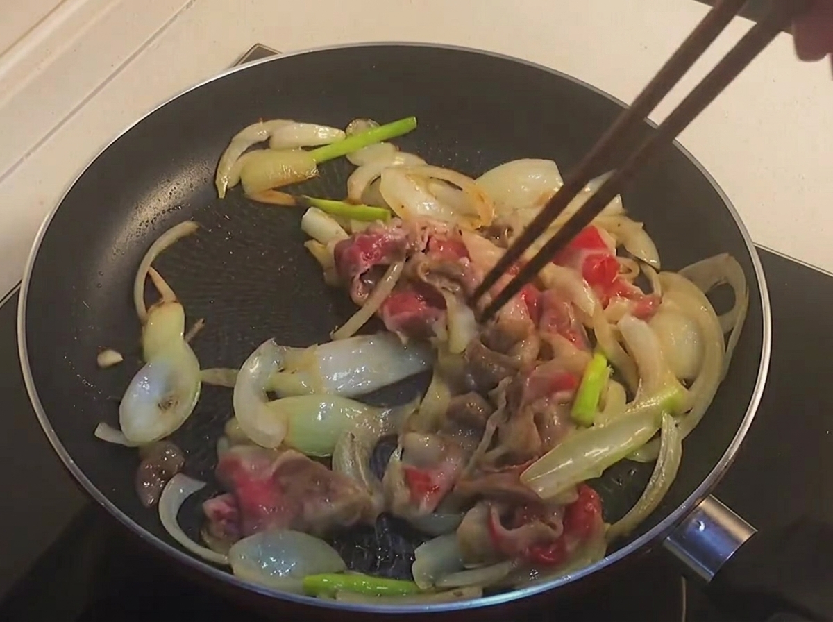 Palillos removiendo rodajas de panceta de ternera parcialmente cocidas y ligeramente doradas junto con cebollas en una sartén negracite: 1.