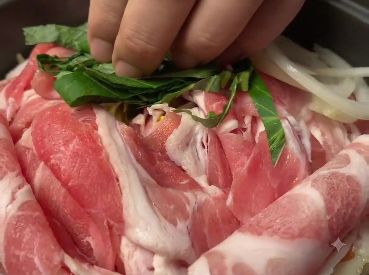 Hojas de perilla verdes picadas siendo esparcidas sobre las rodajas de cerdo crudo en la sartén.