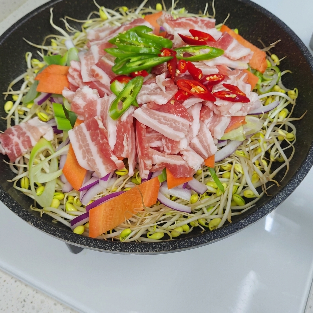 Vista cenital de una sartén llena con una capa de brotes de soja cubierta con cebollas en rodajas, zanahorias, tiras de panceta cruda y rodajas de chile verde y rojo.