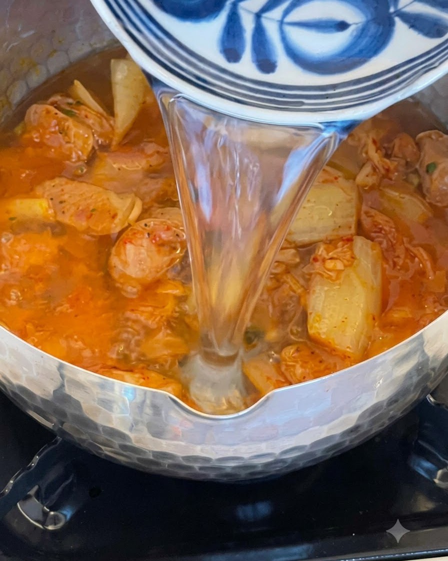 Agua cayendo desde un tazón decorativo azul y blanco hacia una olla llena de kimchi burbujeante de color rojo y caldo de salchicha.