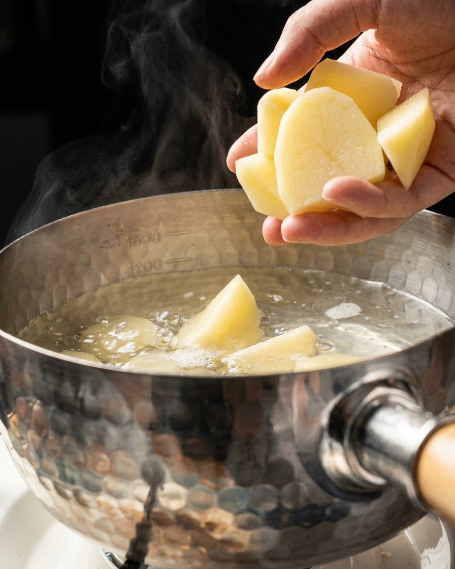 Una mano echando trozos de patata en una olla metálica con agua hirviendo y vapor.