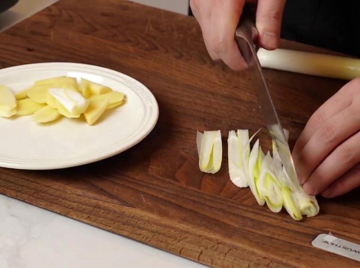 Un chef cortando cebolletas y trozos de jengibre en una tabla de cortar de madera junto a un plato blanco.