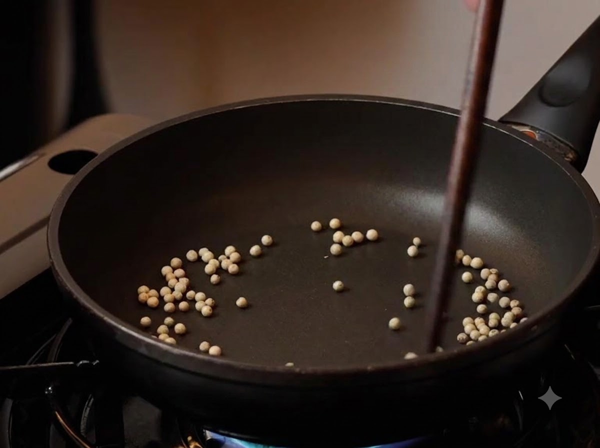 Granos de pimienta blanca tostándose en una sartén negra con palillos removiéndolos a fuego lento.