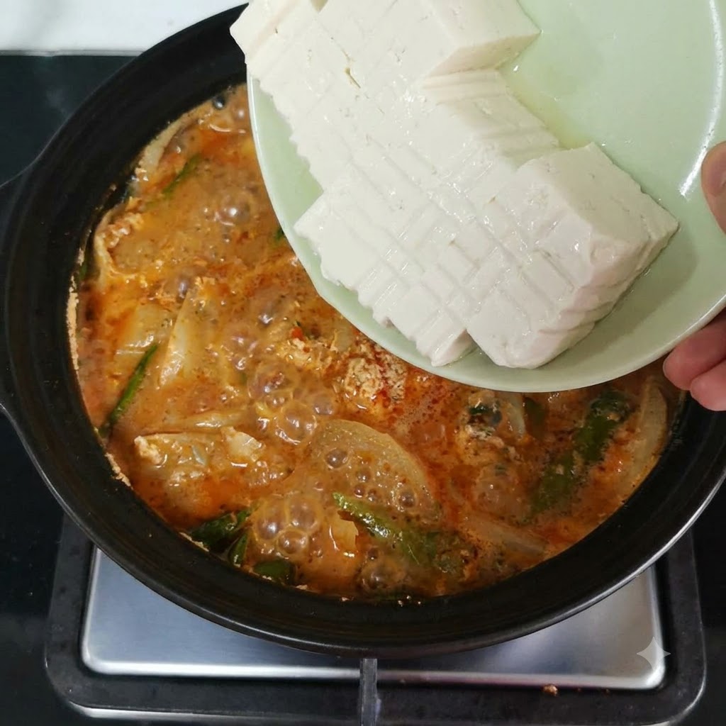 Rodajas de tofu suave vertiéndose desde un plato a una olla a fuego lento llena de caldo rojo anaranjado y verdurascite: 1.
