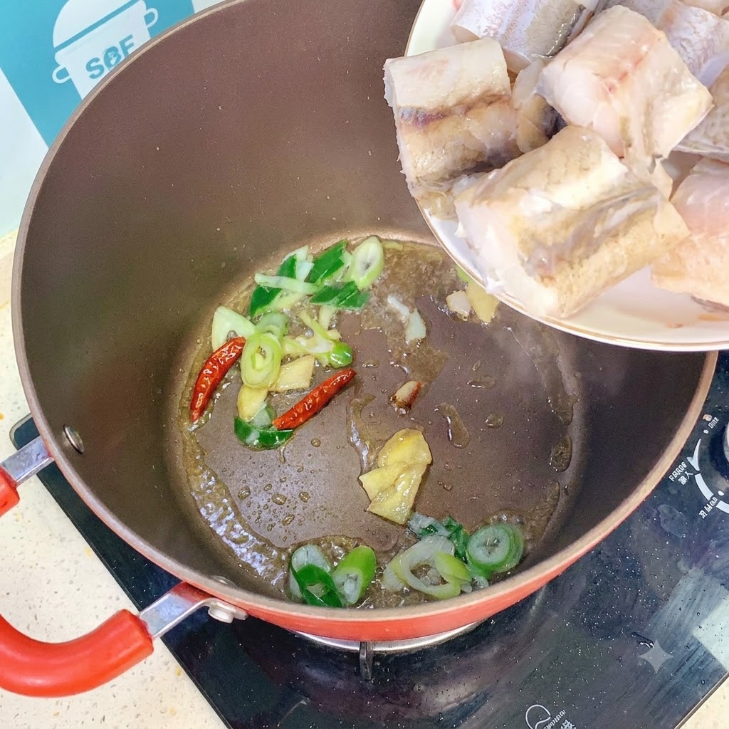 Trozos de abadejo añadiéndose a una olla con cebolletas, jengibre y chiles salteados.