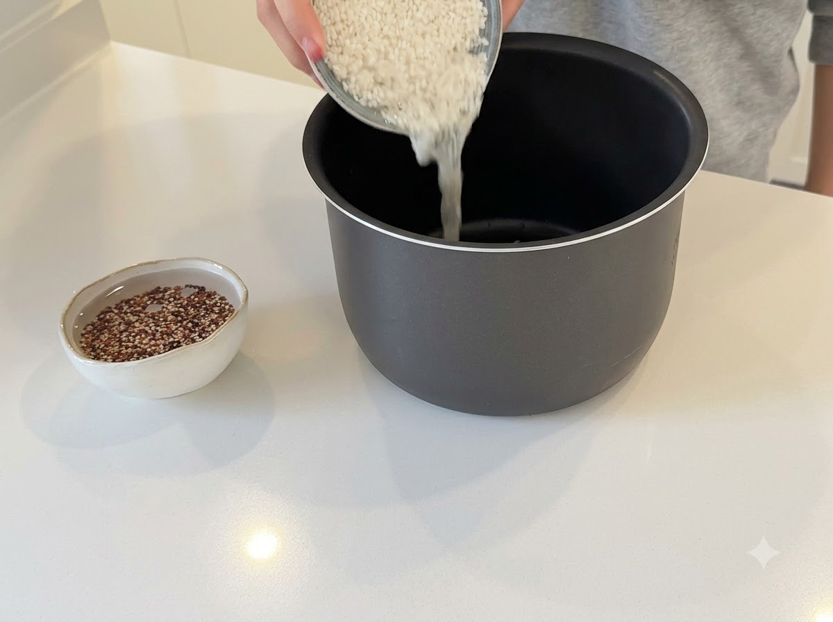 Manos vertiendo arroz blanco en una olla de arrocera oscura con un pequeño bol de quinoa remojándose cerca.