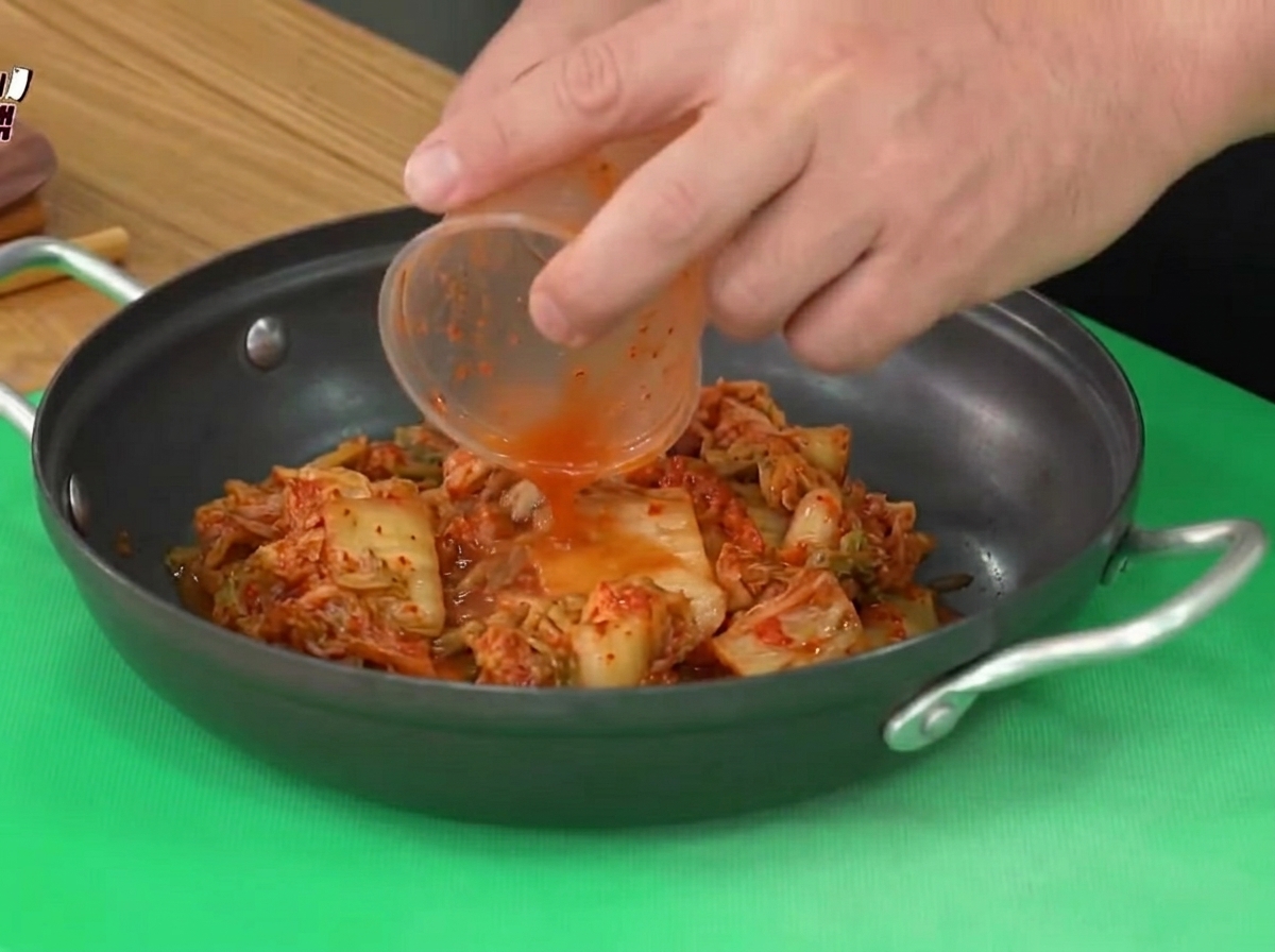 Kimchi agrio siendo agregado desde un vaso de plástico hacia una olla de cocina negra.