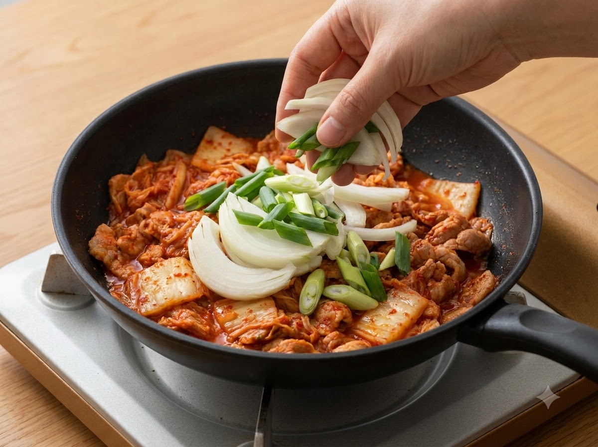 Cebollas cortadas en rodajas y cebollines siendo añadidos a la olla sobre el kimchi.