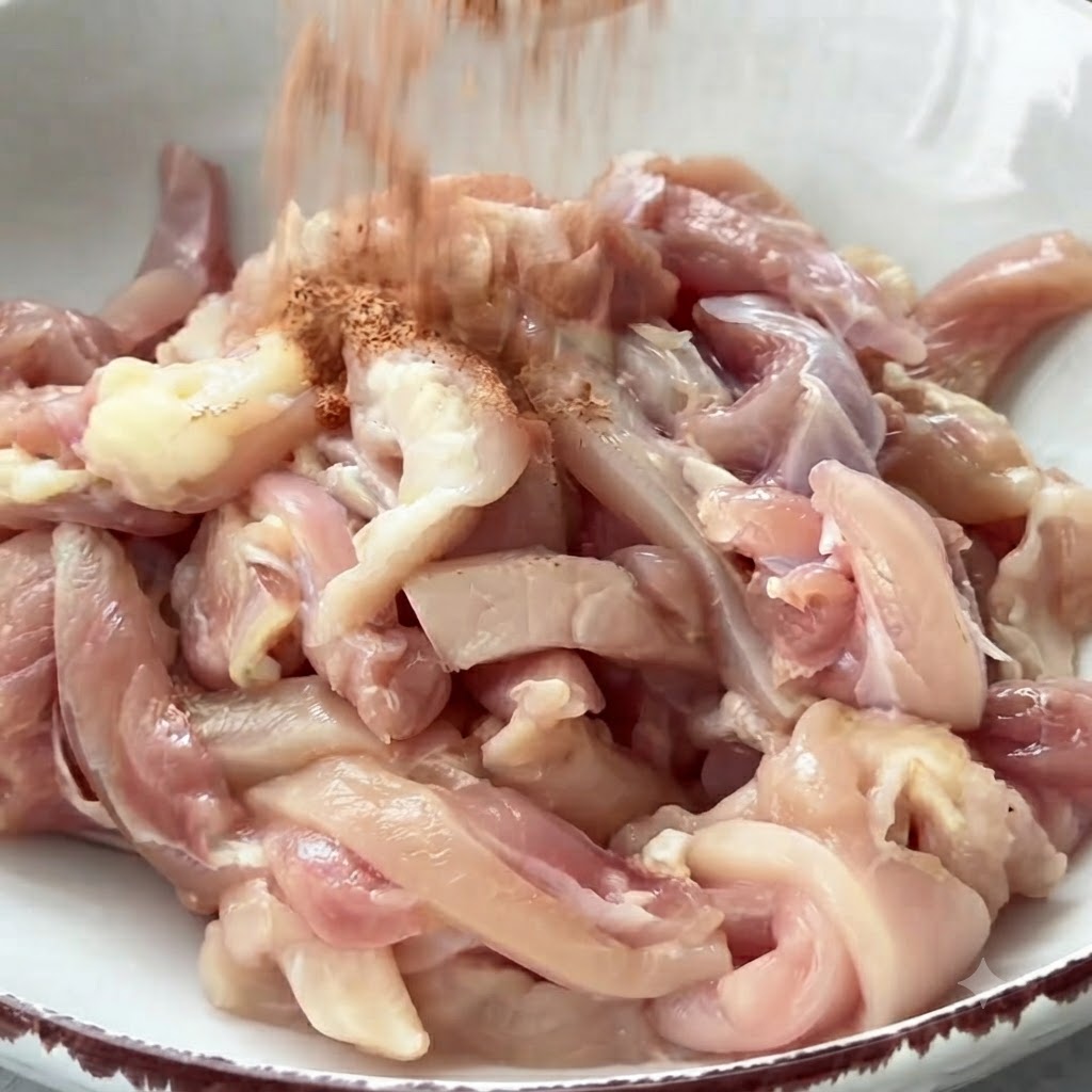 Especias cayendo en forma de lluvia sobre un bol lleno de tiras de pollo crudo listas para ser mezcladas.