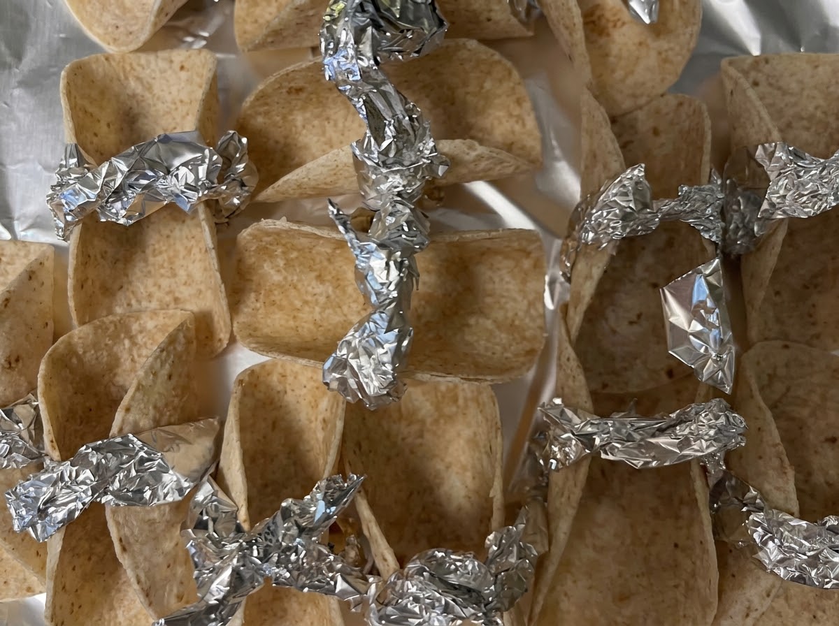Pequeñas envolturas de tortilla sostenidas y aseguradas con trozos de papel de aluminio en una bandeja para hornear listas para el horno.