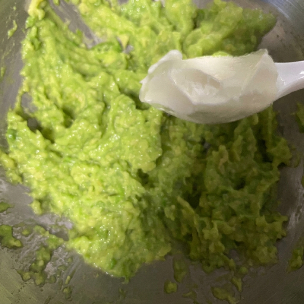 Una cuchara blanca recogiendo yogur griego espeso y agregándolo a un tazón de aguacate recién triturado.