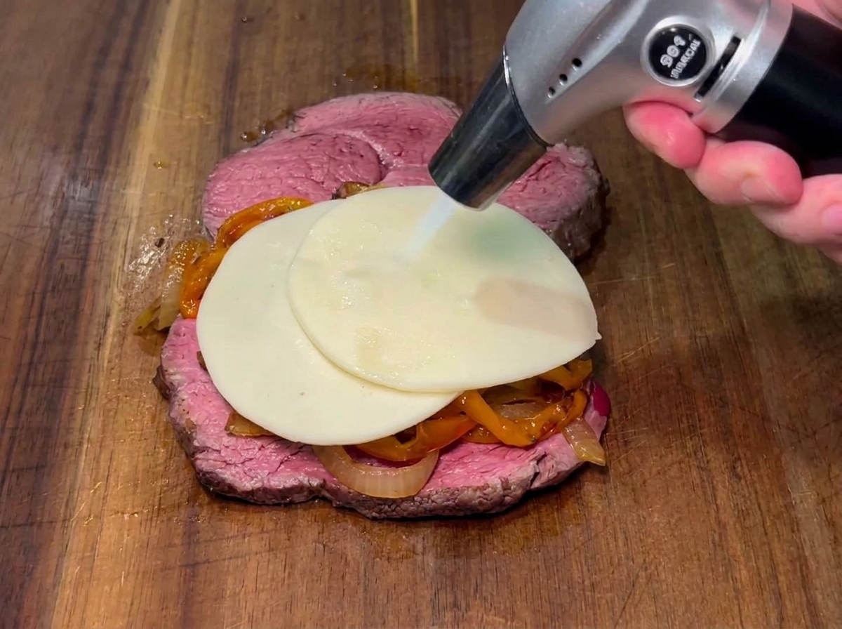 Una rebanada de queso siendo fundida con un soplete de cocina sobre los pimientos y cebollas en un filete.
