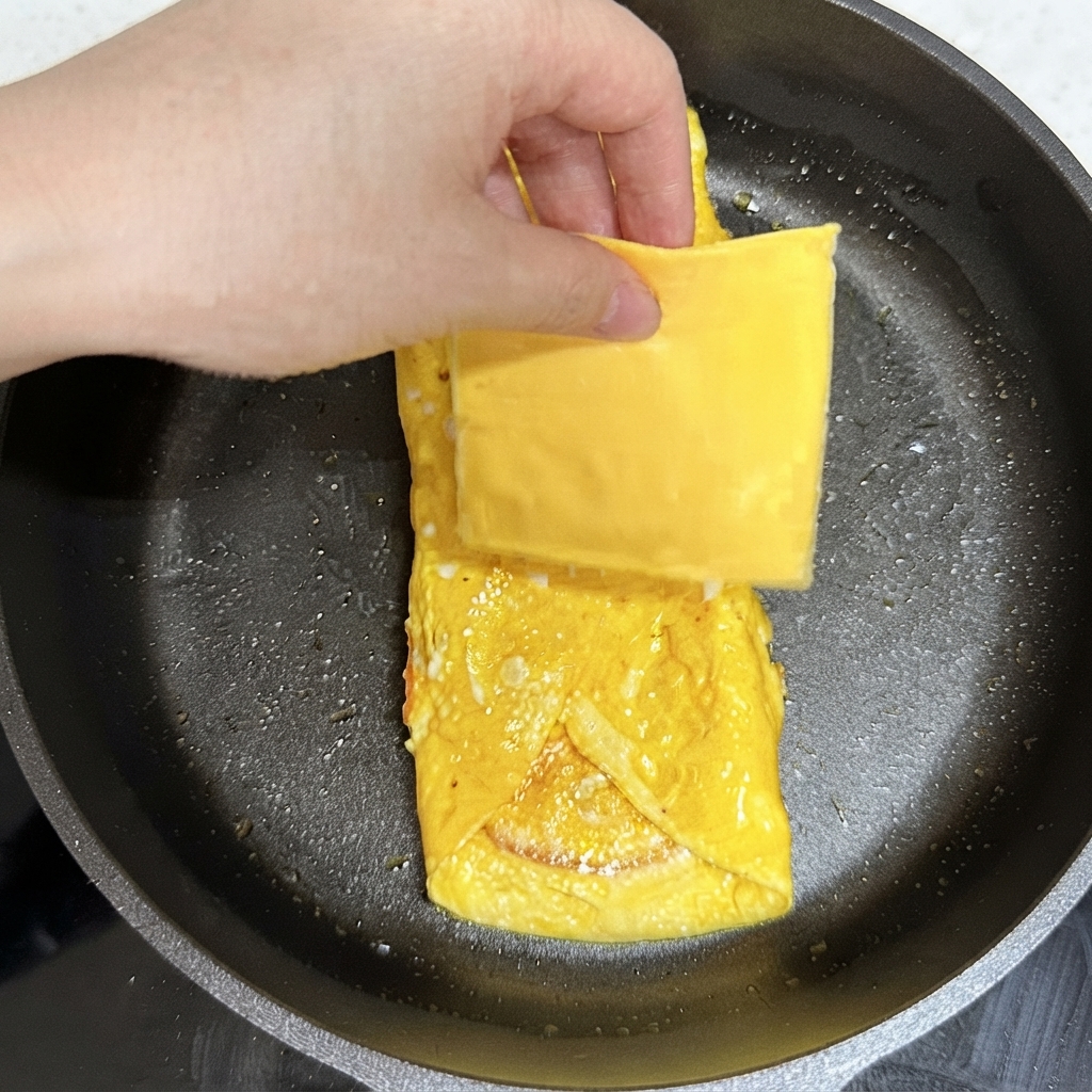 Una mano colocando una loncha cuadrada de queso amarillo sobre el paquete de huevo cocido en la sartén.