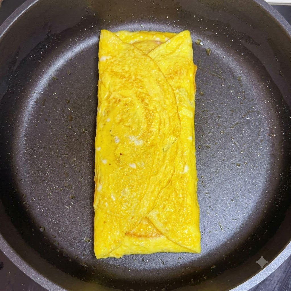 Una loncha doblada de jamón rosado siendo colocada sobre el queso dentro del envoltorio de huevo cocido en la sartén.
