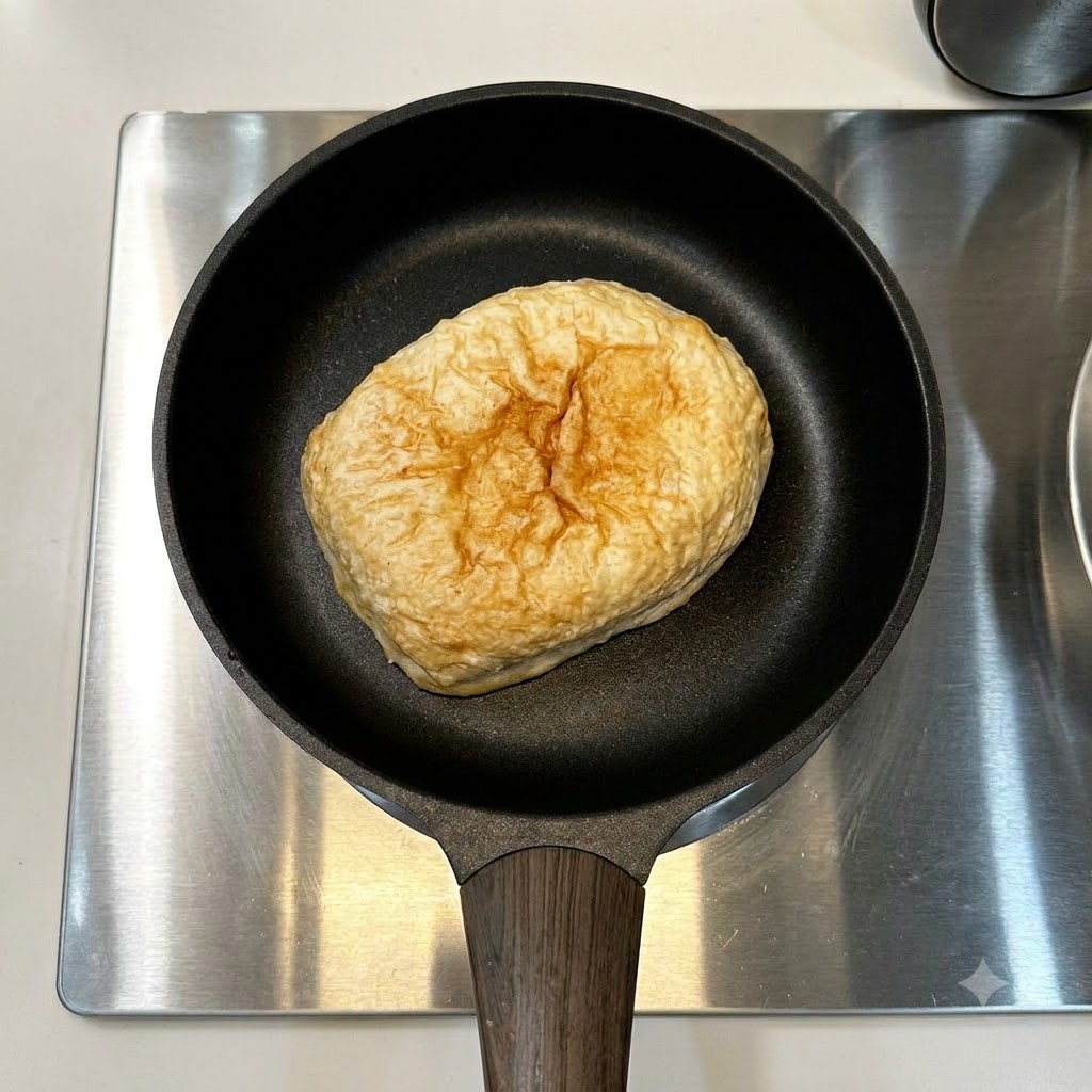 Pan ciabatta y una sola salchicha descansando en una sartén negra sobre una placa de cocina de acero inoxidable.