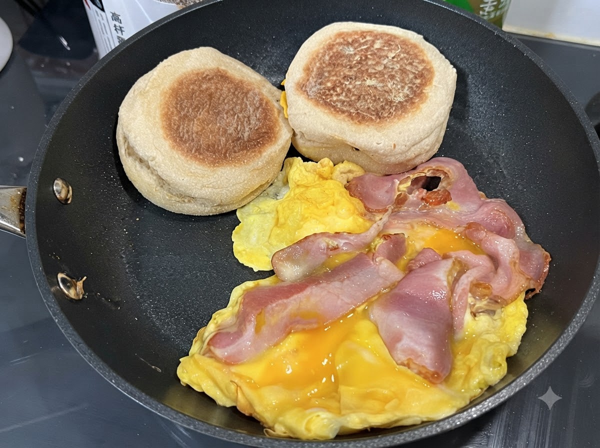 Huevos revolviéndose en una sartén con tiras de tocino y muffins ingleses reposando a un lado.