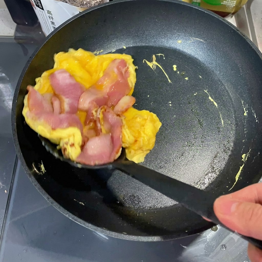 Una espátula levantando una pila de huevo cocido y tocino de una sartén para colocarla sobre la mitad de un muffin inglés tostado.