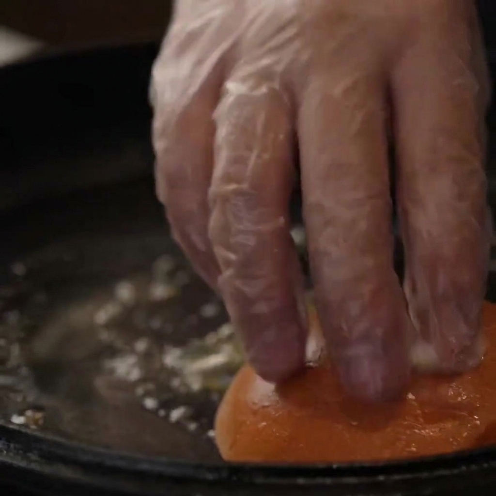 Una mano enguantada presionando los panes de hamburguesa hacia abajo en una sartén de hierro fundido con mantequilla para tostarlos.
