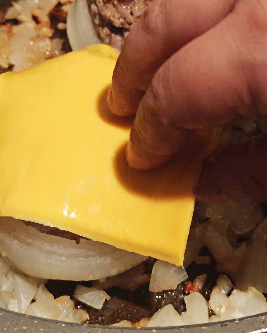 Una loncha de queso amarillo colocándose sobre un aro de cebolla relleno de carne cocinada en la sartén.