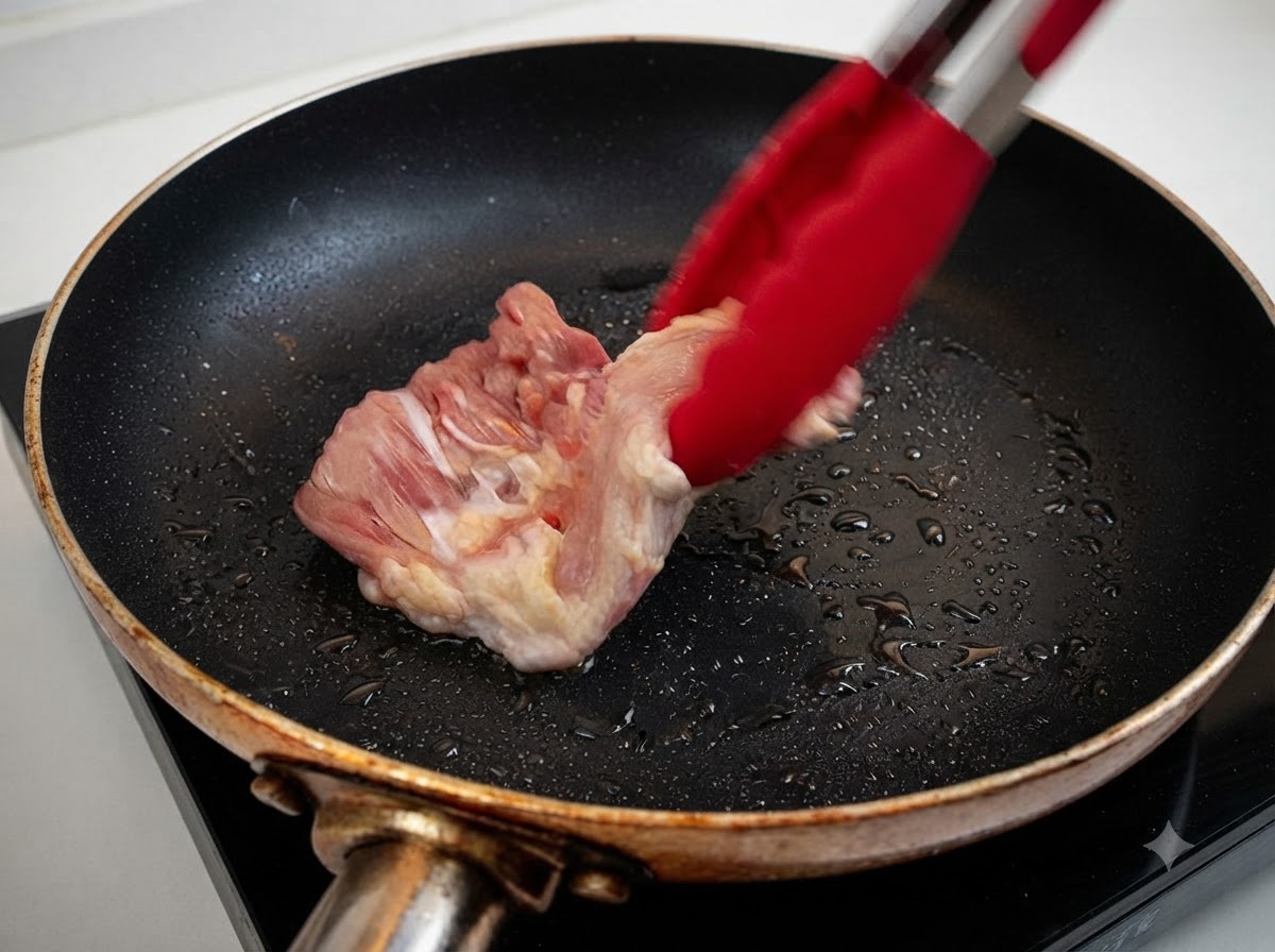 Un muslo de pollo crudo siendo colocado en una sartén caliente con aceite usando pinzas rojas.