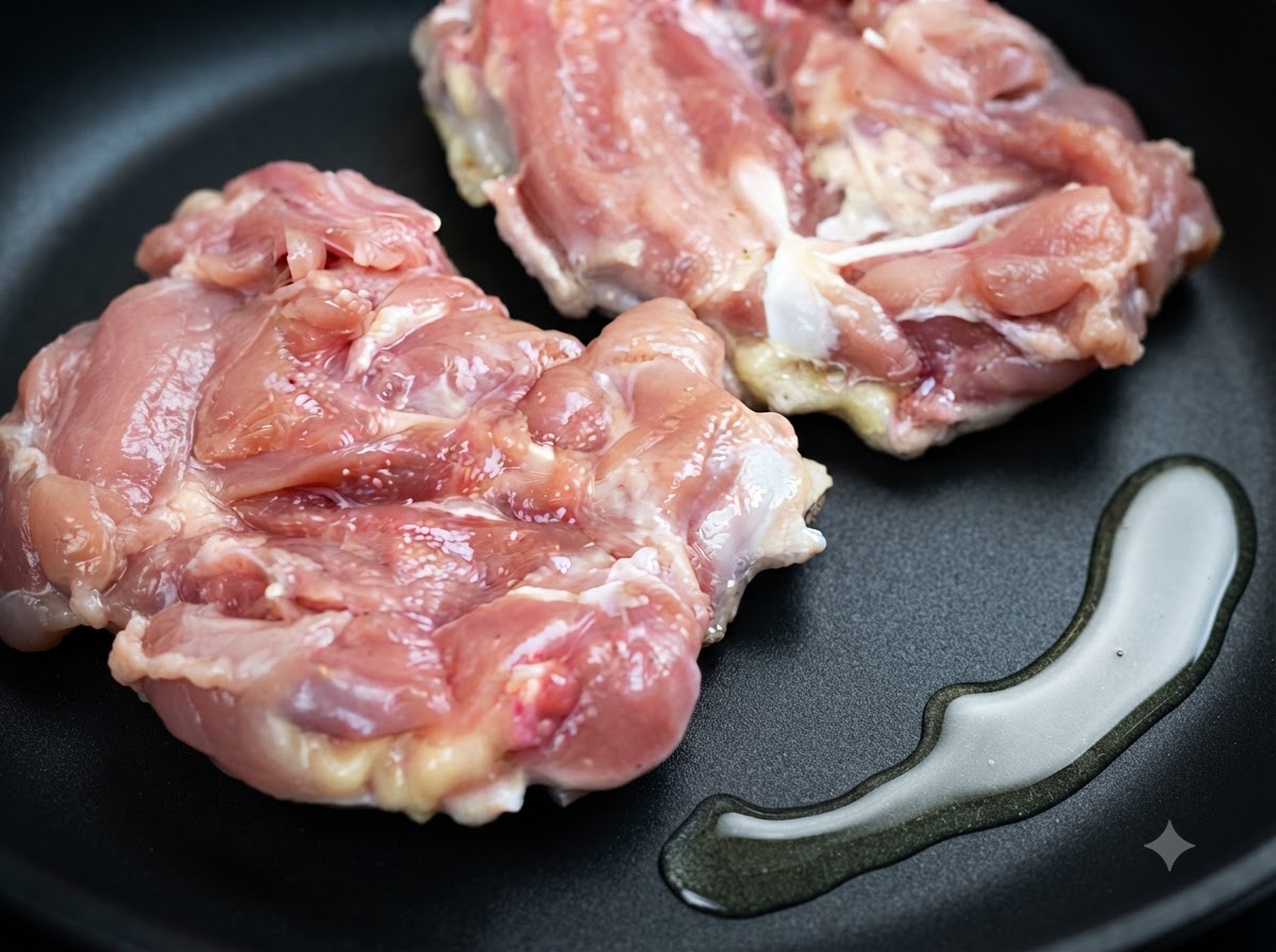 Dos muslos de pollo crudos recostados en una sartén junto a un charco de aceite de cocina.
