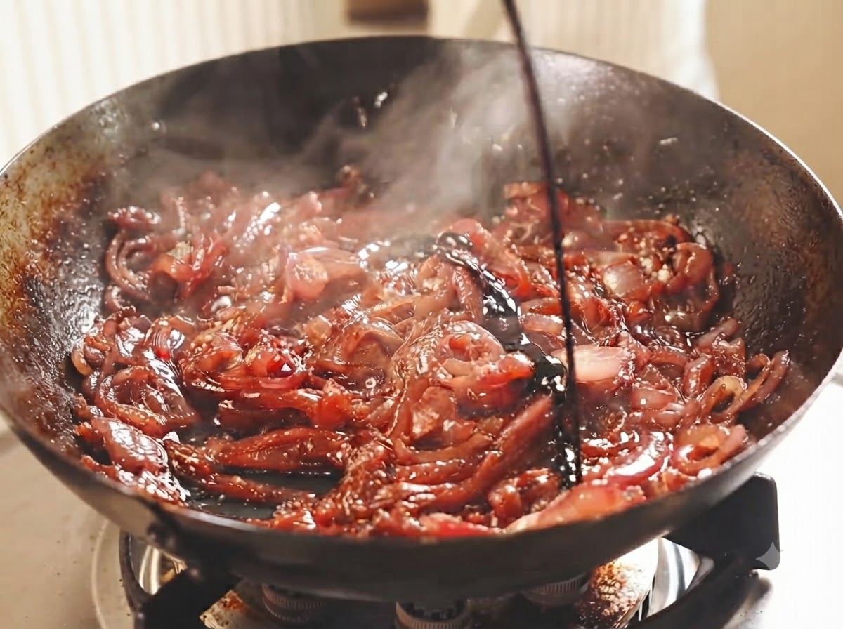 Cebollas rojas cortadas en rodajas cociéndose a fuego lento en un wok oscuro con vino oscuro y vinagre vertidos sobre ellas.