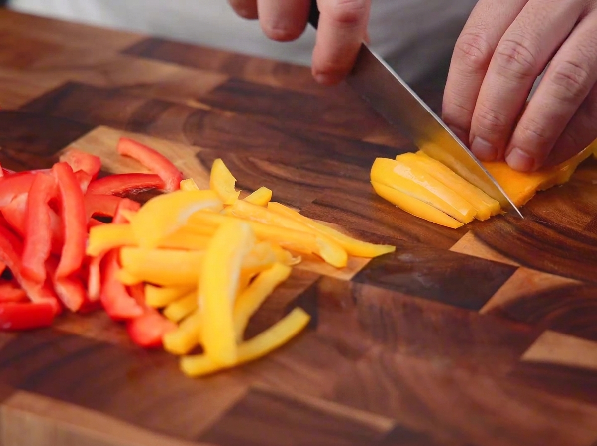 Pimientos rojos y amarillos siendo cortados en tiras finas con un cuchillo sobre una tabla de madera.