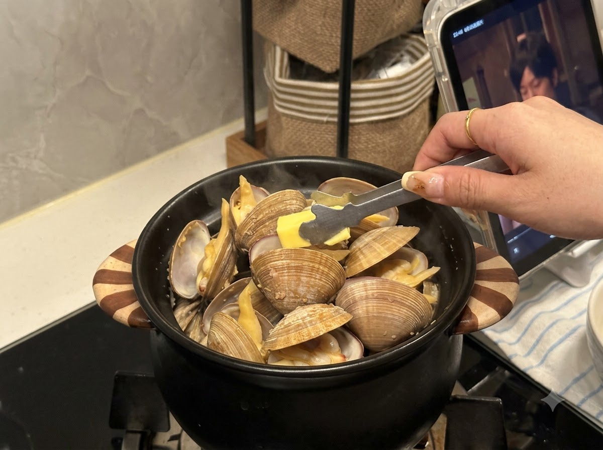 Un pequeño trozo de mantequilla siendo colocado con pinzas en una olla negra llena de almejas abiertas.
