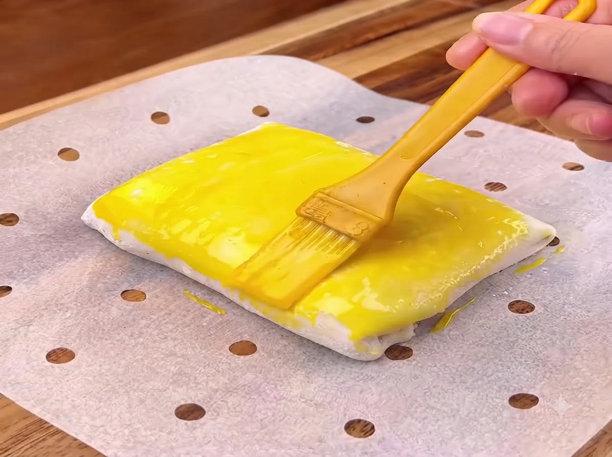 Huevo batido siendo aplicado con un pincel sobre la superficie de un cuadrado de masa doblado.