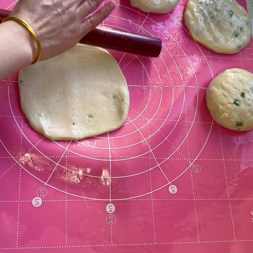 Un rodillo de madera aplanando una porción de masa en un círculo delgado sobre una estera rosa.