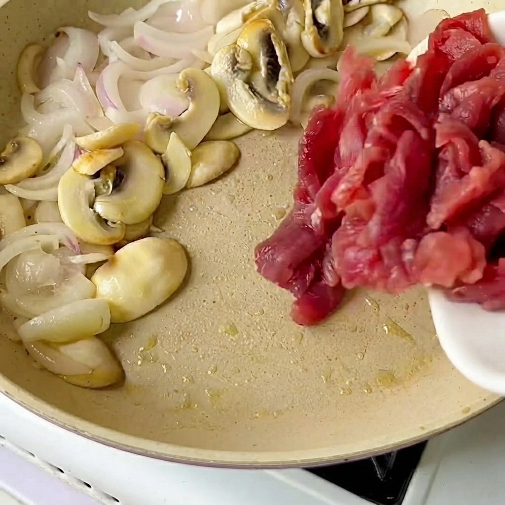 Ternera en rodajas vertida desde un plato blanco a una sartén con cebollas y champiñones salteados.
