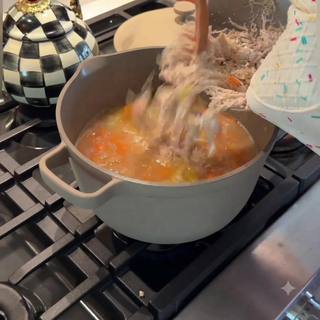 Vertiendo pollo finamente desmenuzado desde un bol de metal de vuelta a una olla gris de sopa de pollo y verduras a fuego lento.