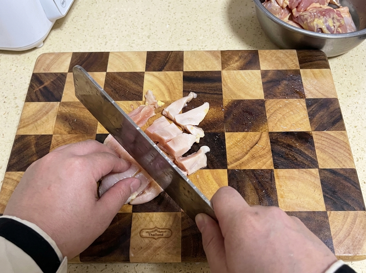 Un cuchillo de carnicero cortando pechuga de pollo cruda en trozos pequeños sobre una tabla de cortar de madera a cuadros.