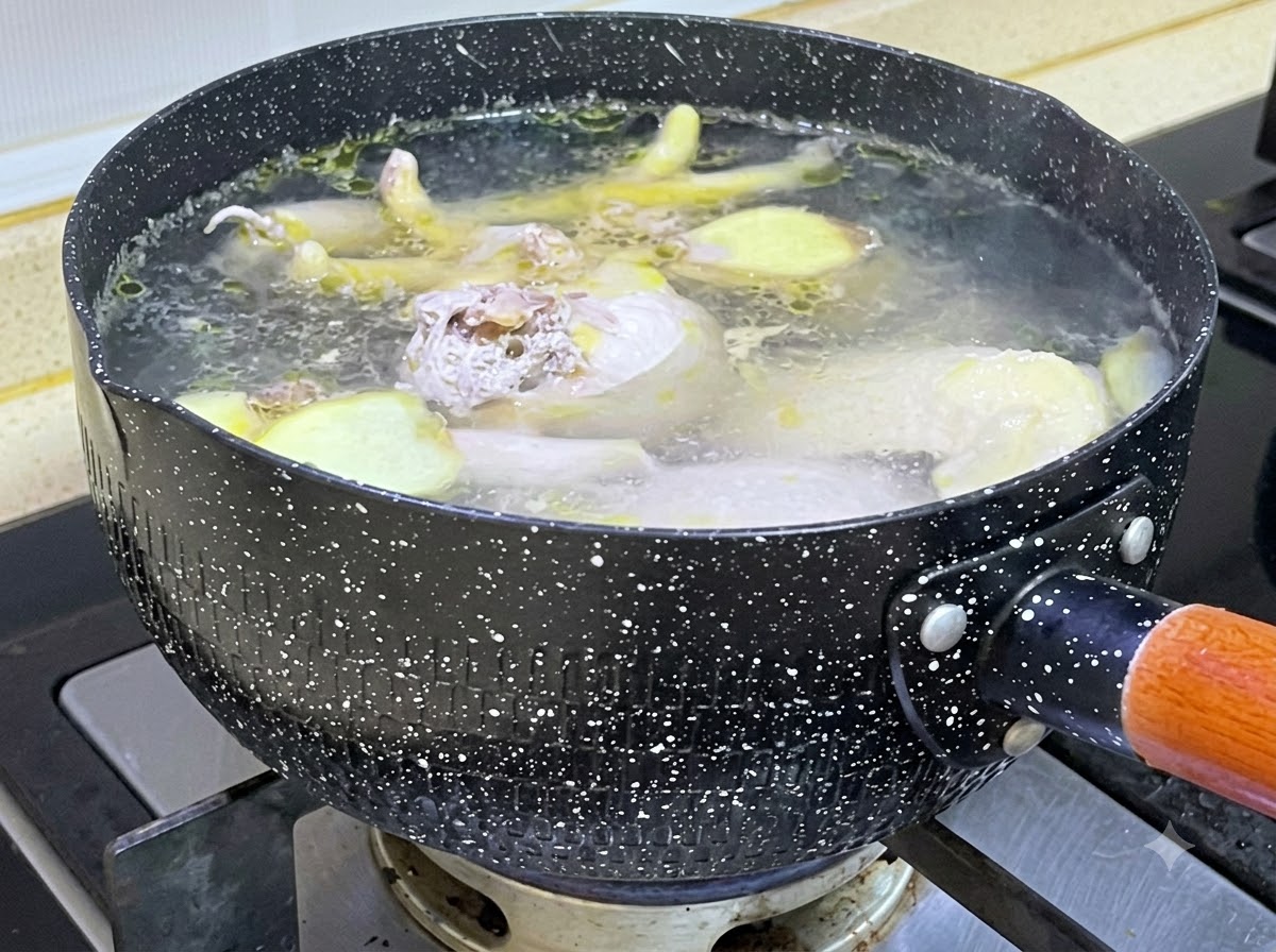 Trozos de pollo crudo y rodajas de jengibre hirviendo a fuego lento en una olla negra con motas llena de agua hirviendo.