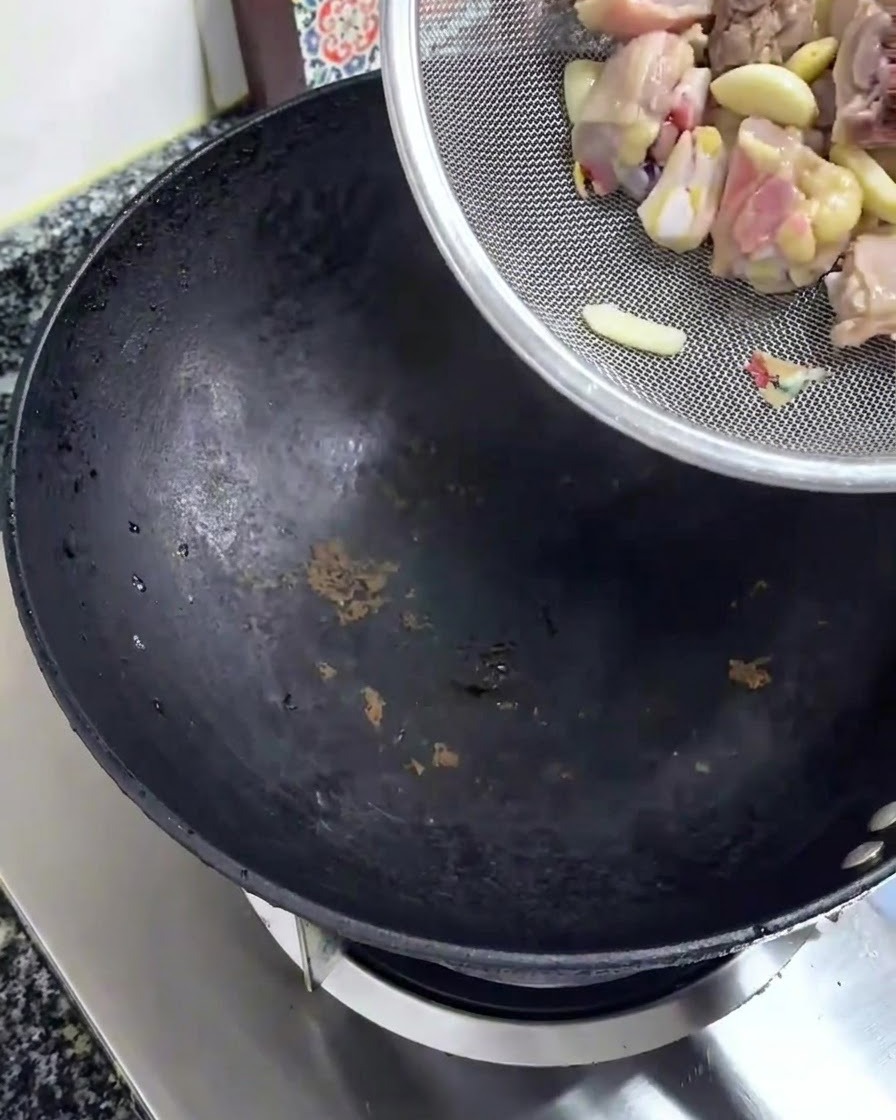 Trozos de pollo crudo cayendo desde un colador plateado a un wok negro.