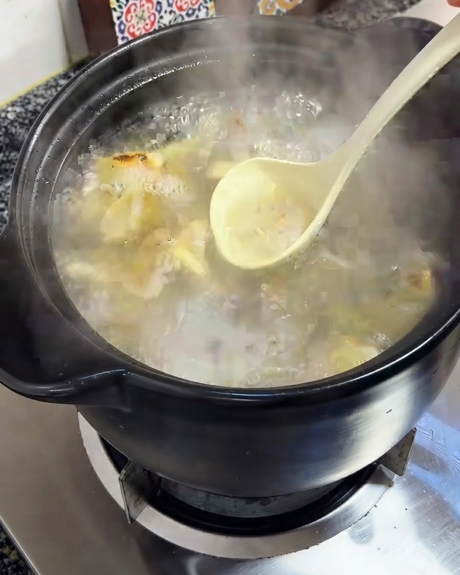 Un cucharón quitando la espuma de la superficie del caldo de pollo burbujeante en una olla de barro negra.